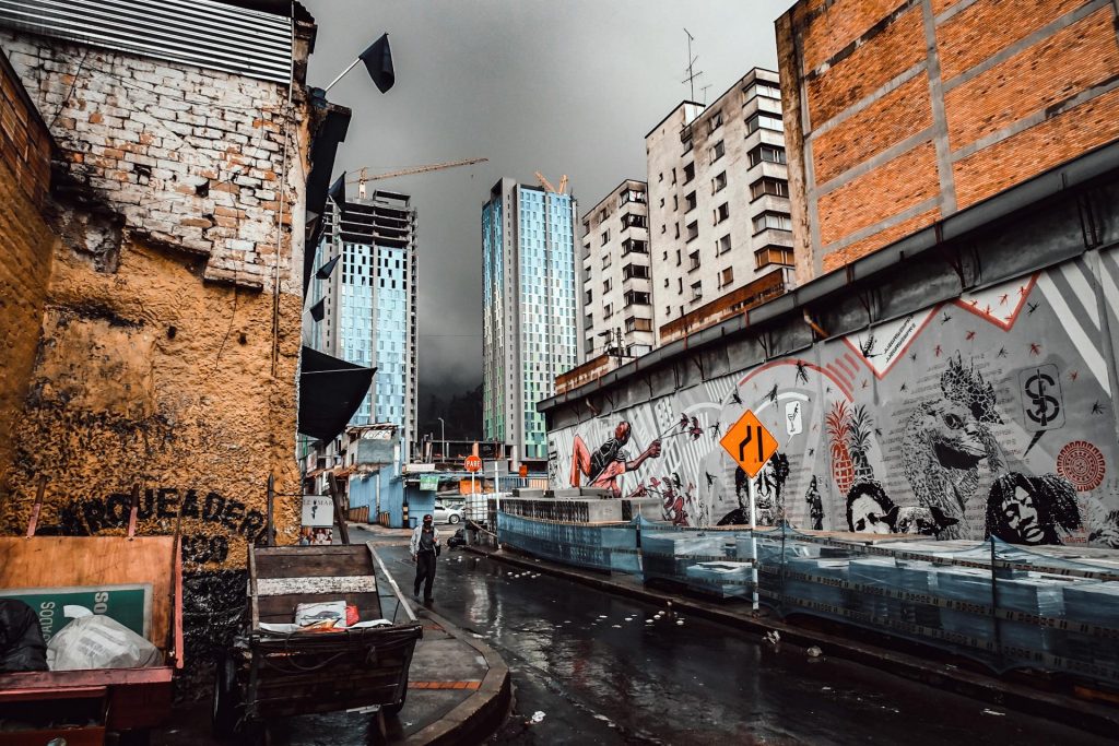 Person Walking On A Wet Street Along A Building With Mural Under Gray Sky in Bogotá, Colombia