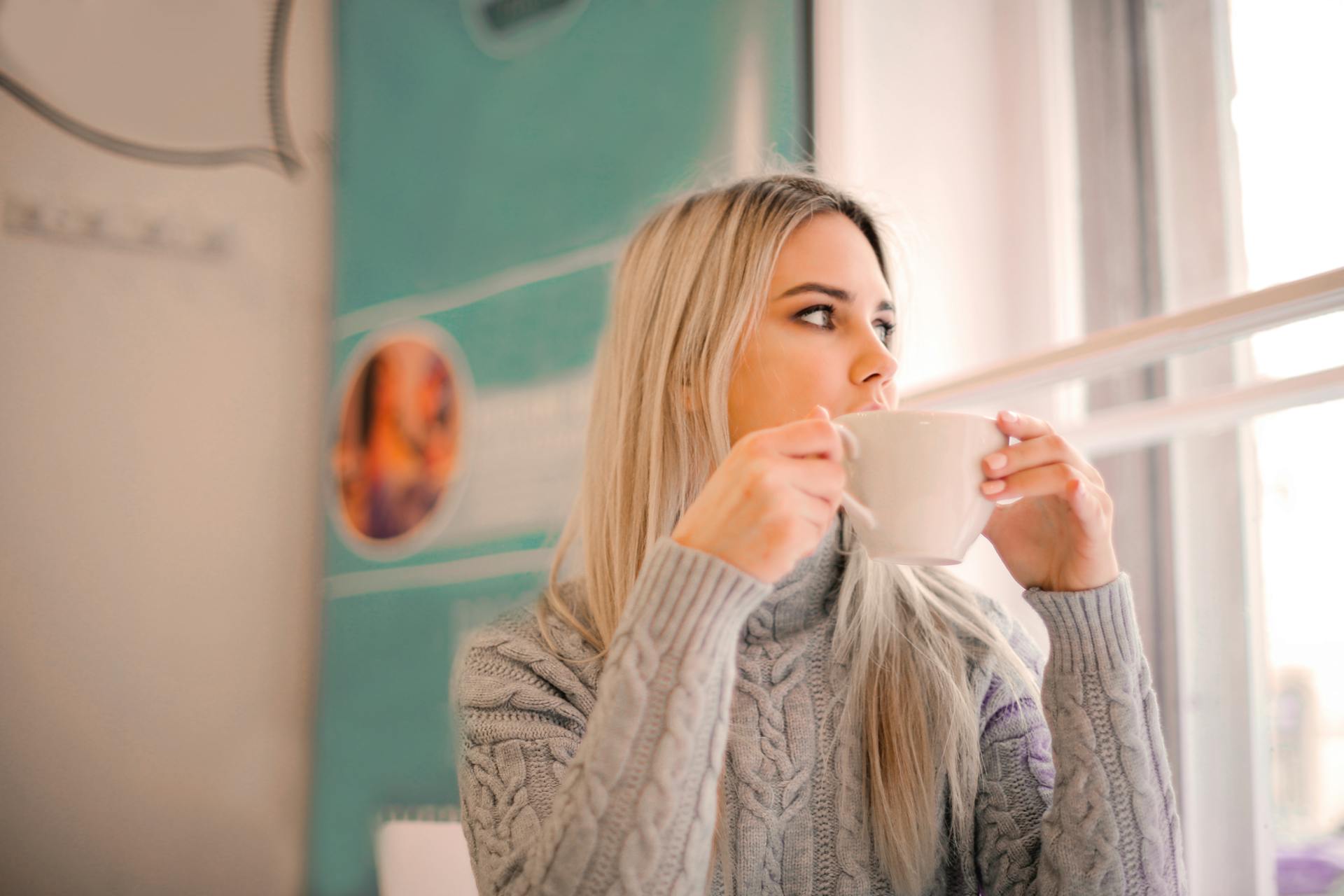 Woman in Gray Sweater Drinking from White Ceramic Mug While looking Away