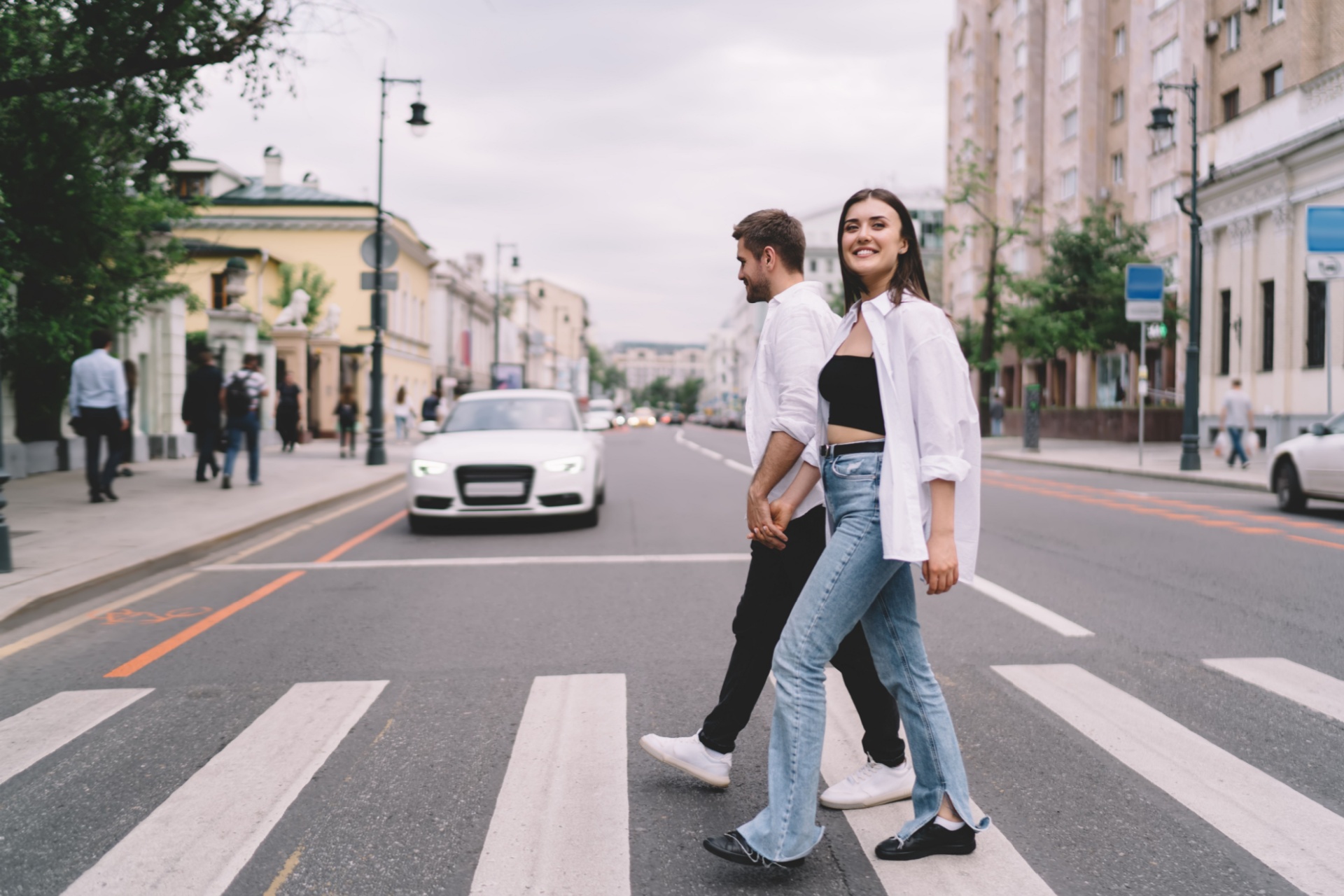 Full body of happy young couple in casual clothing holding hands while crossing road on crosswalk in Moscow town during daytime