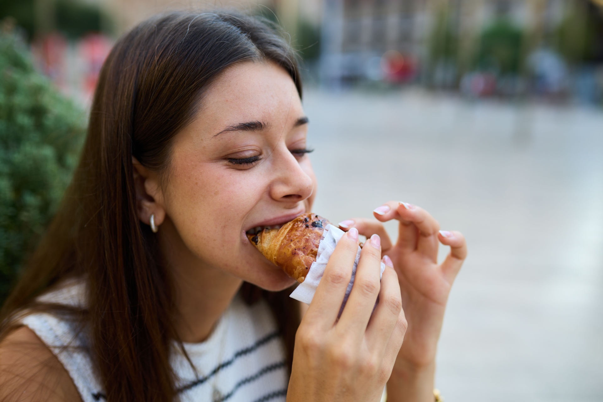 Happy woman eating a pastry croissant, smiling and looking away on a city street