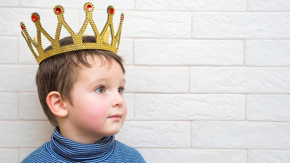 Little child with a crown on his head against a white brick wall