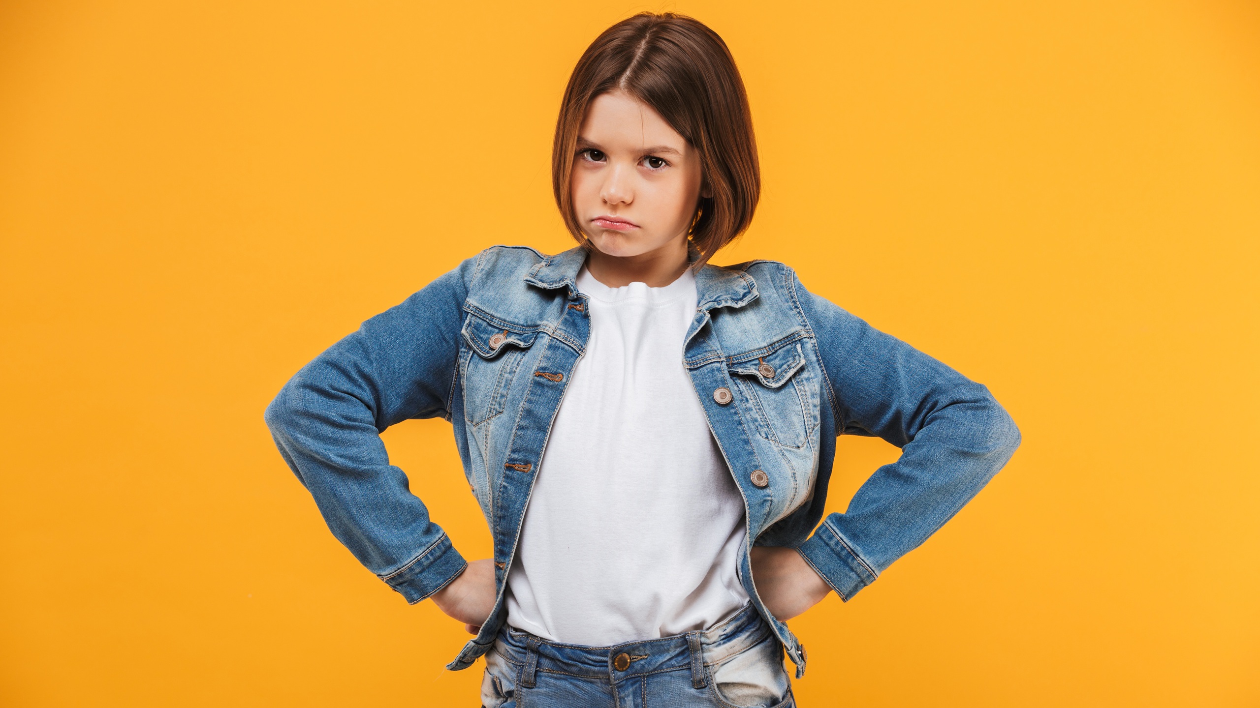 Portrait of an upset little schoolgirl standing with hands on hips over yellow background