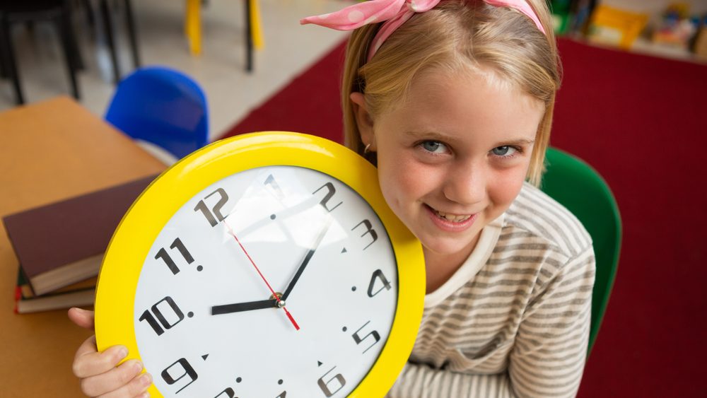 Close-up of a Caucasian smiling schoolgirl holding a wall clock and looking at the camera in the classroom