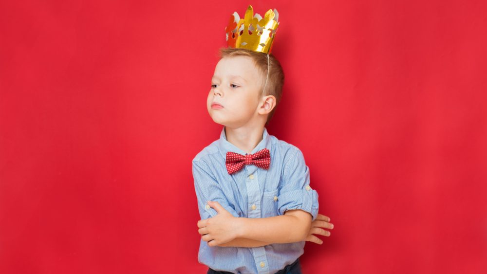 Happy education and childhood concepts with an adorable 6-year old boy holding a golden king crown on his head as a wise spoiled child, red background