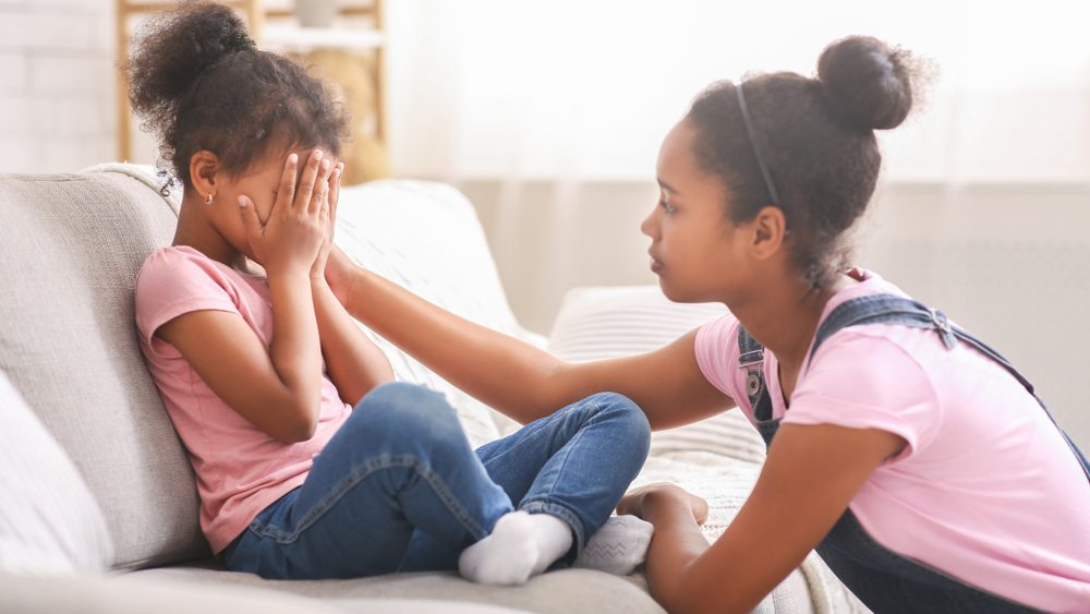 Caring african american teen girl calming her sad crying baby sister, side view