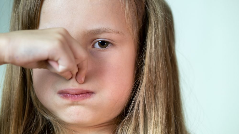 Close-up portrait of little girl with long hair holding her nose with fingers in disgust.