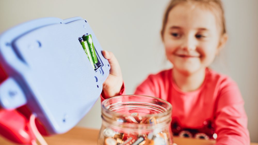 Little girl removing used batteries from toy and putting into jar for recycling. Child separating waste. Batteries only container