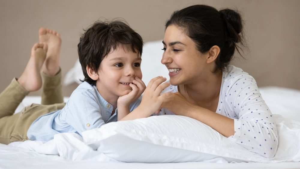 Happy Indian little boy and cheerful mom resting in bed, talking, laughing, having fun, enjoying leisure together in weekend morning. Motherhood, time with family, comfortable house concept