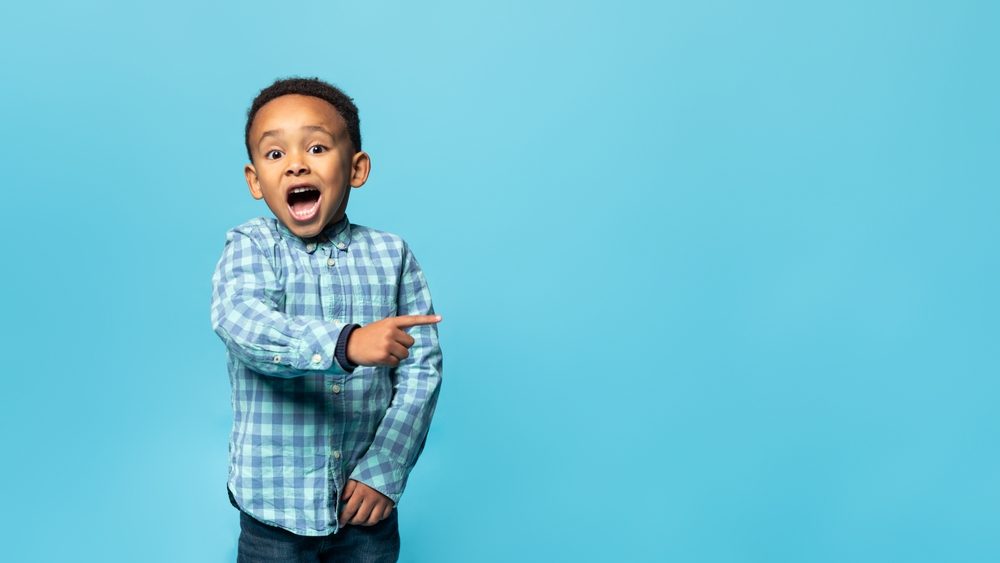 Special offer. Adorable african american boy pointing his finger aside at copy space and looking at camera with open mouth, posing over blue studio wall, banner, panorama