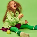 Emotionally eating fruits. Little cute girl, child with curly hair posing with apples over green studio background. Concept of childhood, emotions, fun, fashion, lifestyle, facial expression