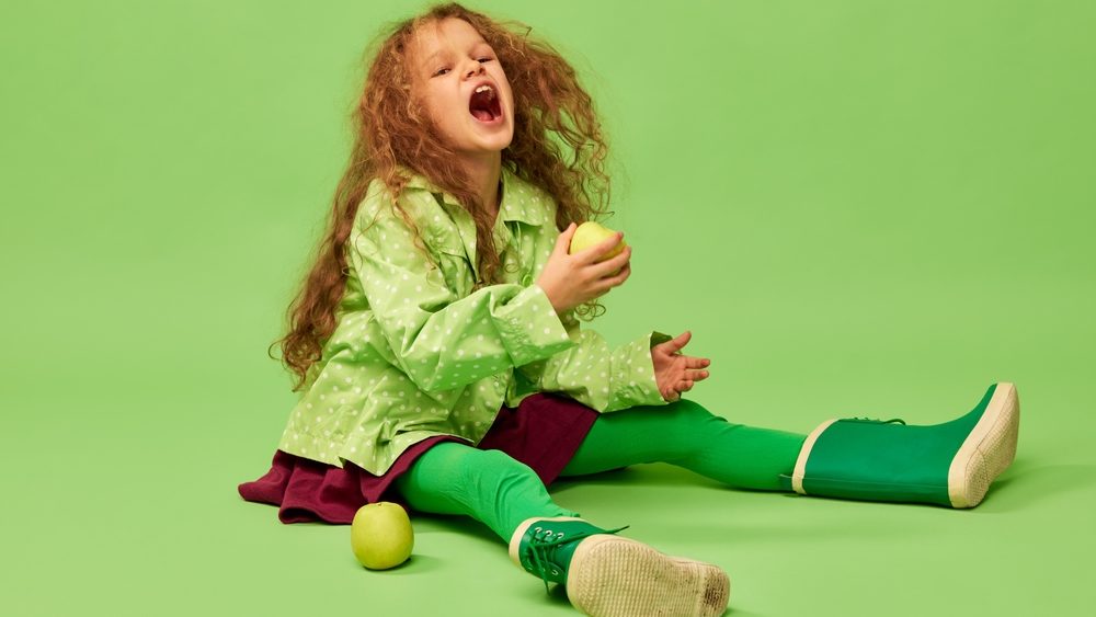 Emotionally eating fruits. Little cute girl, child with curly hair posing with apples over green studio background. Concept of childhood, emotions, fun, fashion, lifestyle, facial expression