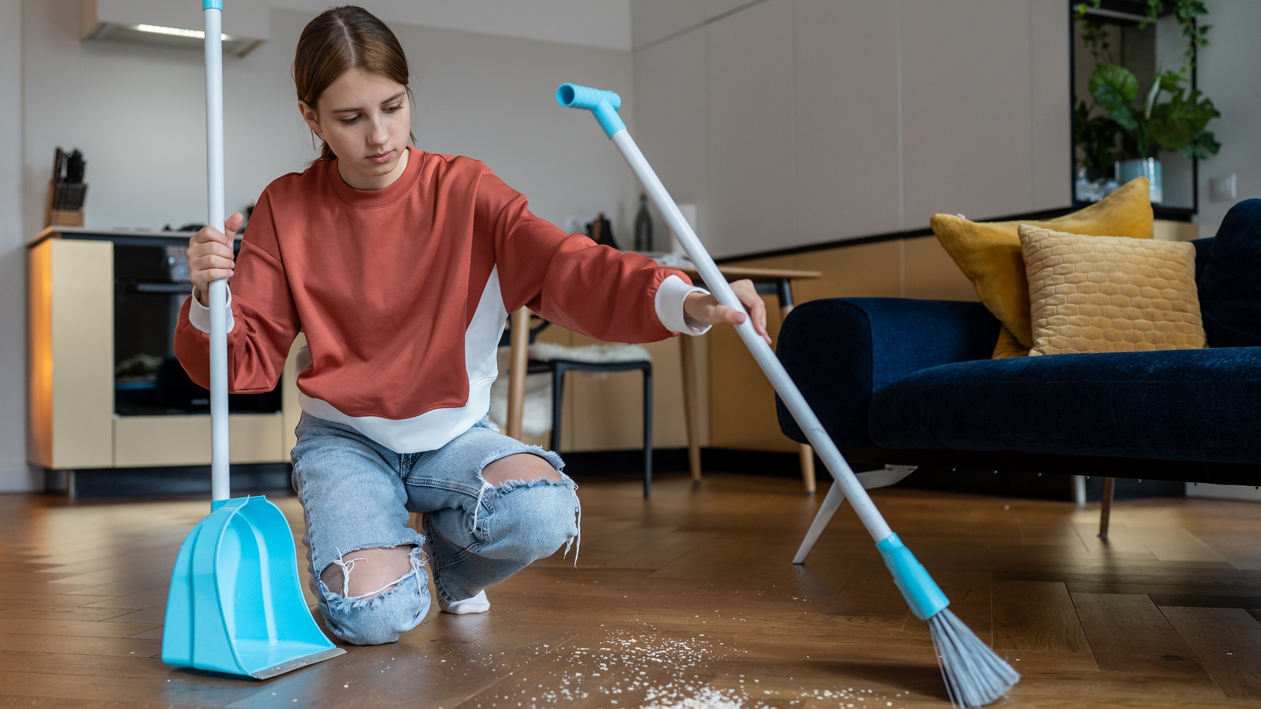 Independent teenager. Habit of cleaning house since childhood. Focused teen girl sweeps floor from scattered debris with broom with brush and dustpan. Adolescent do house chores, domestic cleanup.
