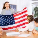 Group of curious preteen learners and young female teacher with flag of USA in classroom of academy