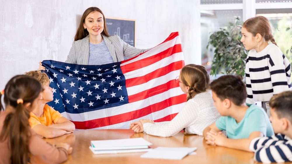 Group of curious preteen learners and young female teacher with flag of USA in classroom of academy