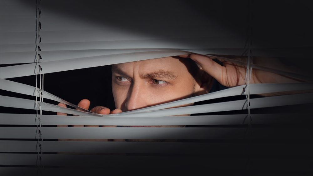 Worried man looking through window blinds into darkness. Paranoia concept