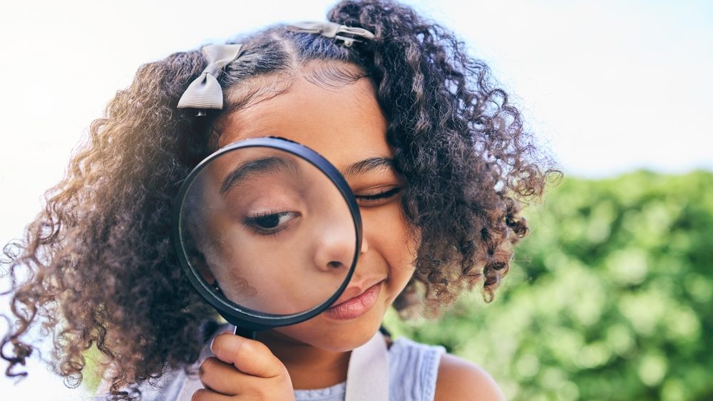Girl child, magnifying glass and inspection in garden, backyard or park in science, study or outdoor. Young female kid, lens and zoom for nature, search or check for plant, leaves or growth in summer