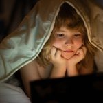 Kid watching tablet before sleeping. Child reads the e-book. Little boy looks at the screen of the tablet at hight. Kid boy playing tablet lying on a bed under blanket. Child social media addiction.