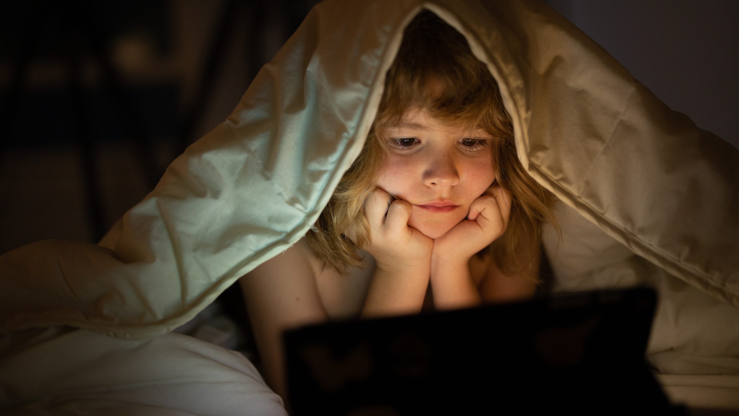 Kid watching tablet before sleeping. Child reads the e-book. Little boy looks at the screen of the tablet at hight. Kid boy playing tablet lying on a bed under blanket. Child social media addiction.