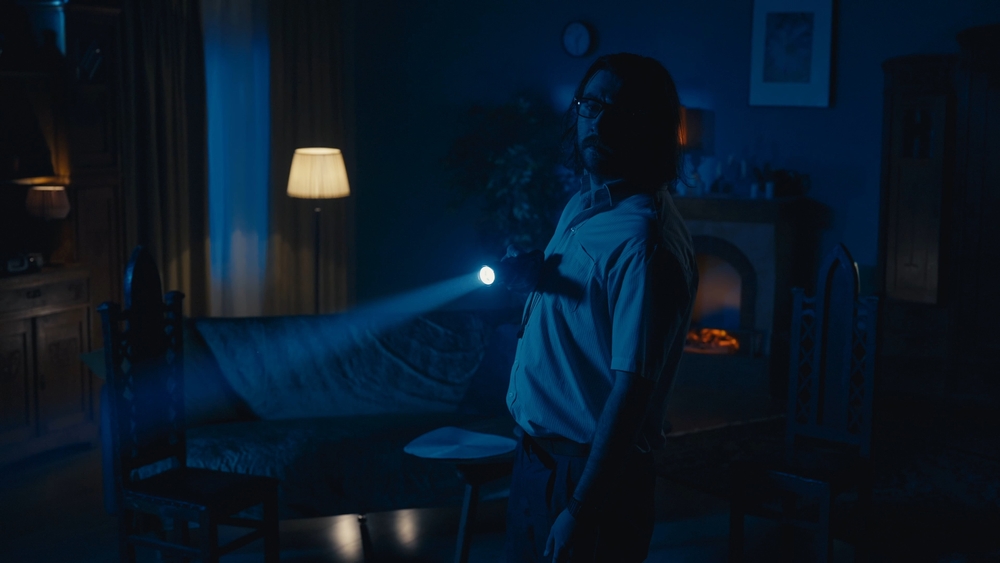 Medium-full photo capturing a man walking around a dark room in search for something strange or paranormal, navigating in space using flashlight.