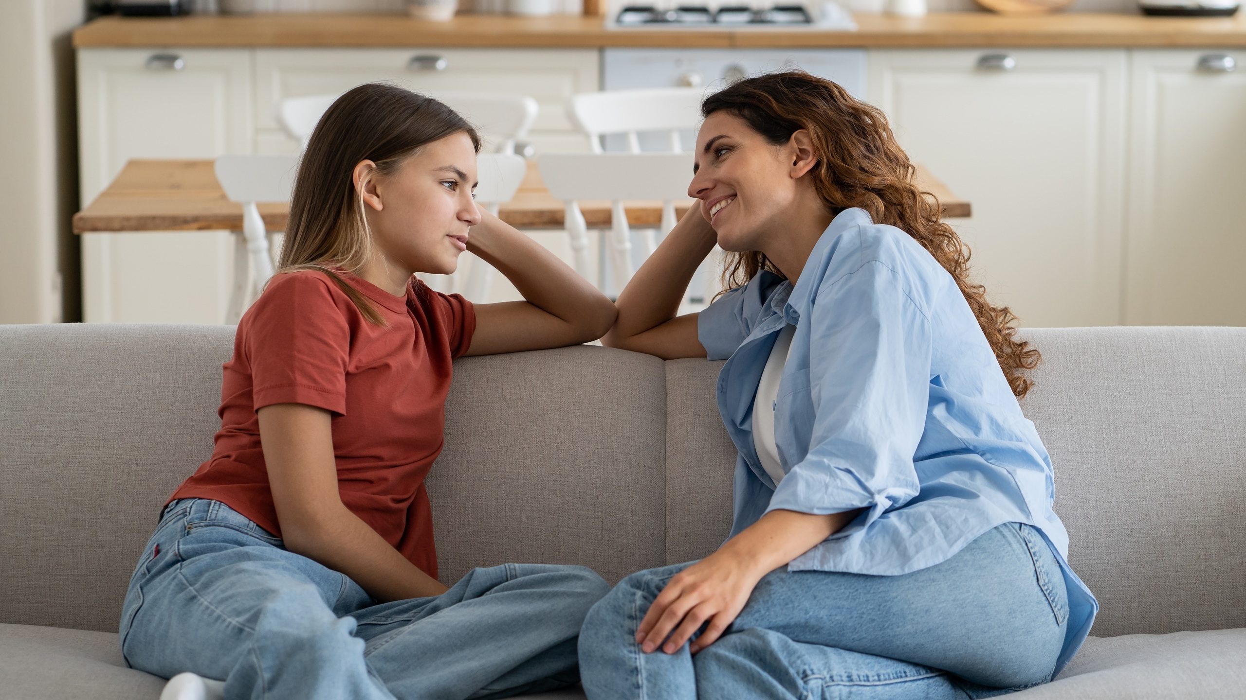 Happy charismatic Italian woman talking with daughter about school friends and summer vacation plans sits on sofa in home interior. Shy casual teen girl shares secrets with mom thanks to family trust 