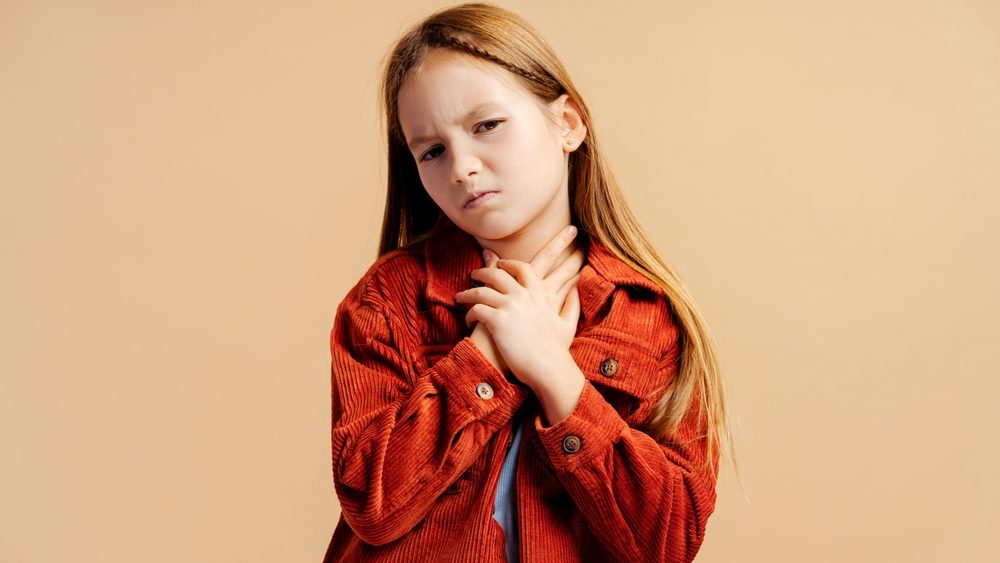 Portrait of sick little girl wearing casual jacket has sore throat looking at camera standing isolated on beige background. Treatment concept 