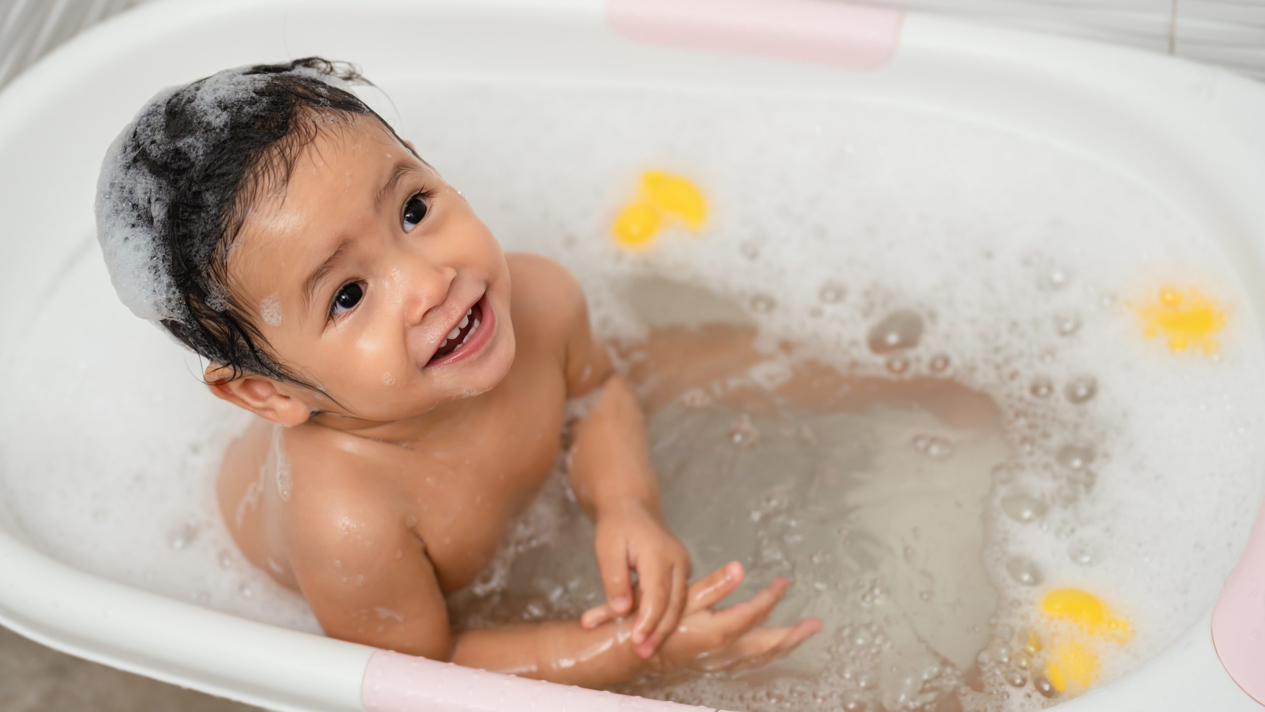 happy infant baby take a bath and playing with foam bubbles in bathtub at home