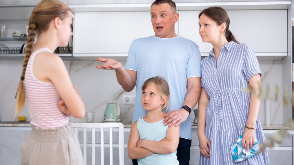 Displeased mother and father reprimanding eldest teenage daughter while standing with her younger sister in home kitchen. Conflict between parents and children