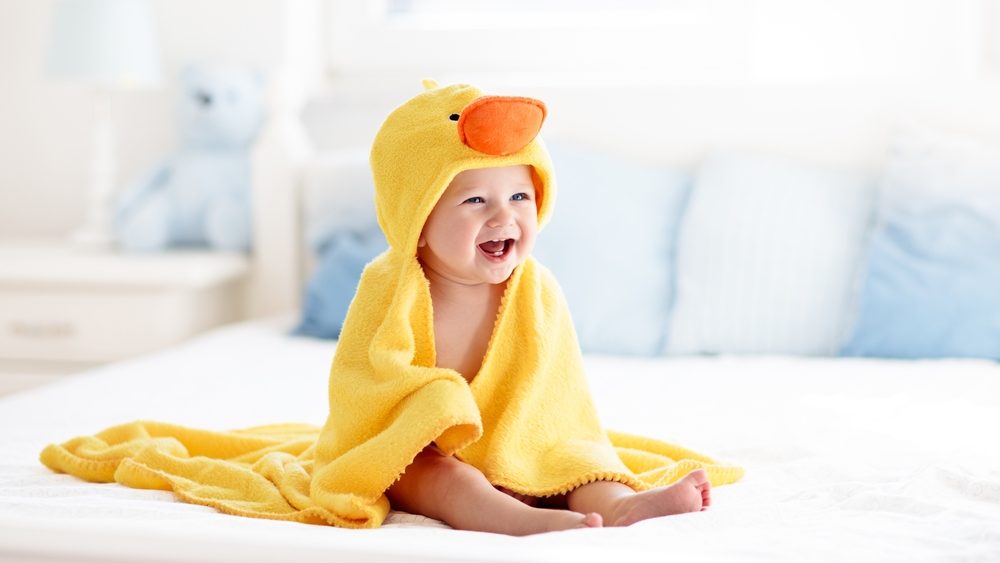 Happy laughing baby wearing yellow hooded duck towel sitting on parents bed after bath or shower. Clean dry child in bedroom. Bathing and washing of little kids. Children hygiene. Textile for infants.