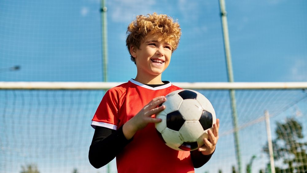 A young boy stands in front of a soccer goal, holding a soccer ball with a determined expression. He is positioned for a kick, showcasing his love for the sport and his readiness to score a goal.