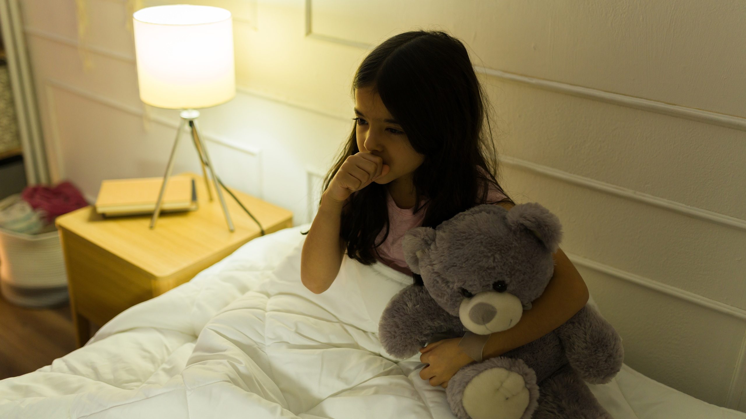 Hispanic little girl holding a teddy bear in her bedroom at home and coughing and feeling unwell
