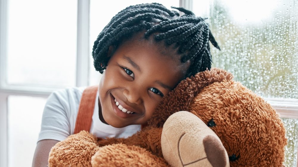 Black girl, portrait and teddy bear in house by window for happy, wellness and playing with toys. Homeless child, smile and stuffed animal in orphanage or children home for love, friendship and hug