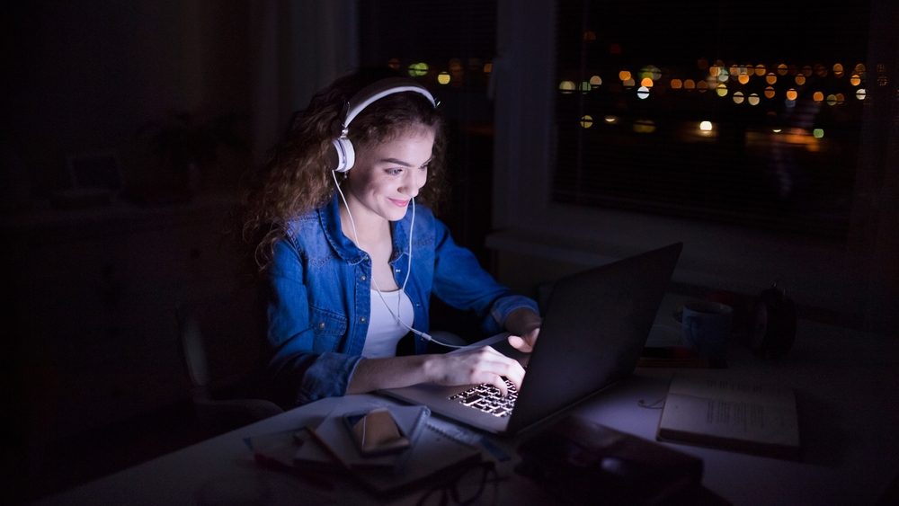 Beautiful university student is writing her thesis on a laptop at night, sitting at home at table, focusing on her research. Preparing for final exam, studying.