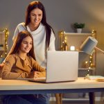 Young cheerful happy mother with her teen school girl daughter studying online class on laptop at home during remote lesson. Woman parent helping child with homework. Distance education.
