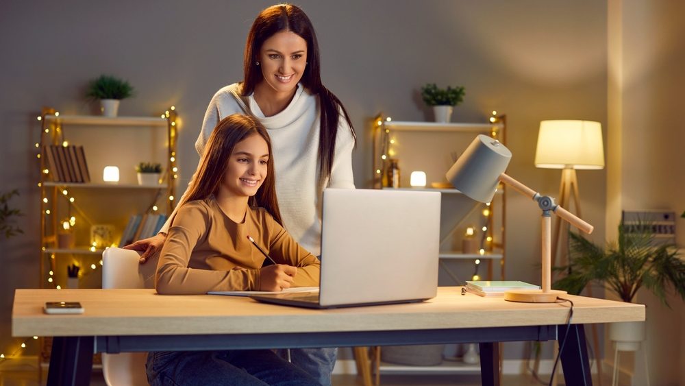 Young cheerful happy mother with her teen school girl daughter studying online class on laptop at home during remote lesson. Woman parent helping child with homework. Distance education.