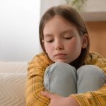 Stressed unhappy sad pensive Caucasian kid little girl feeling frustrated depressed lonely hugging knees alone child bored anxious punished daughter thinking family abuse problems home couch apartment