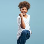 Excited, happy African American boy with curly hairstyle wearing white t shirt, winner posing isolated on blue background. Victory concept