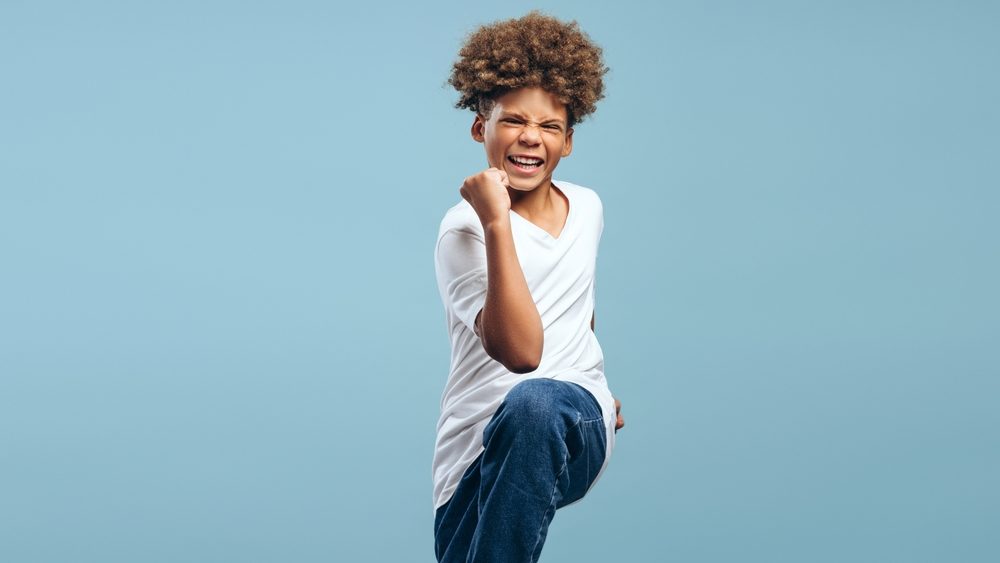 Excited, happy African American boy with curly hairstyle wearing white t shirt, winner posing isolated on blue background. Victory concept