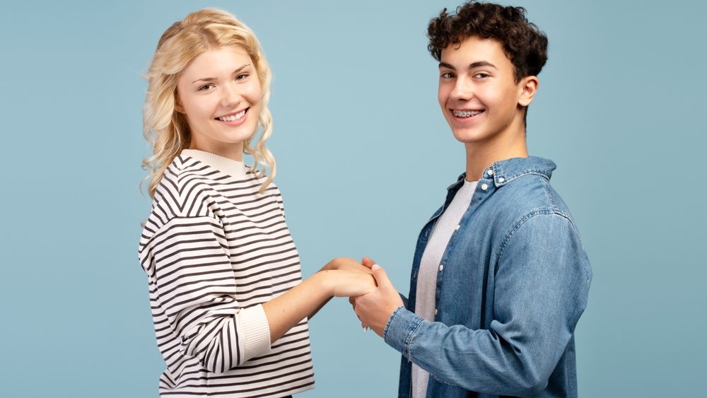 Happy, attractive young couple, smiling boy and girl wearing casual clothes holding hands each other, looking at camera, together standing isolated on blue background. Relationship concept