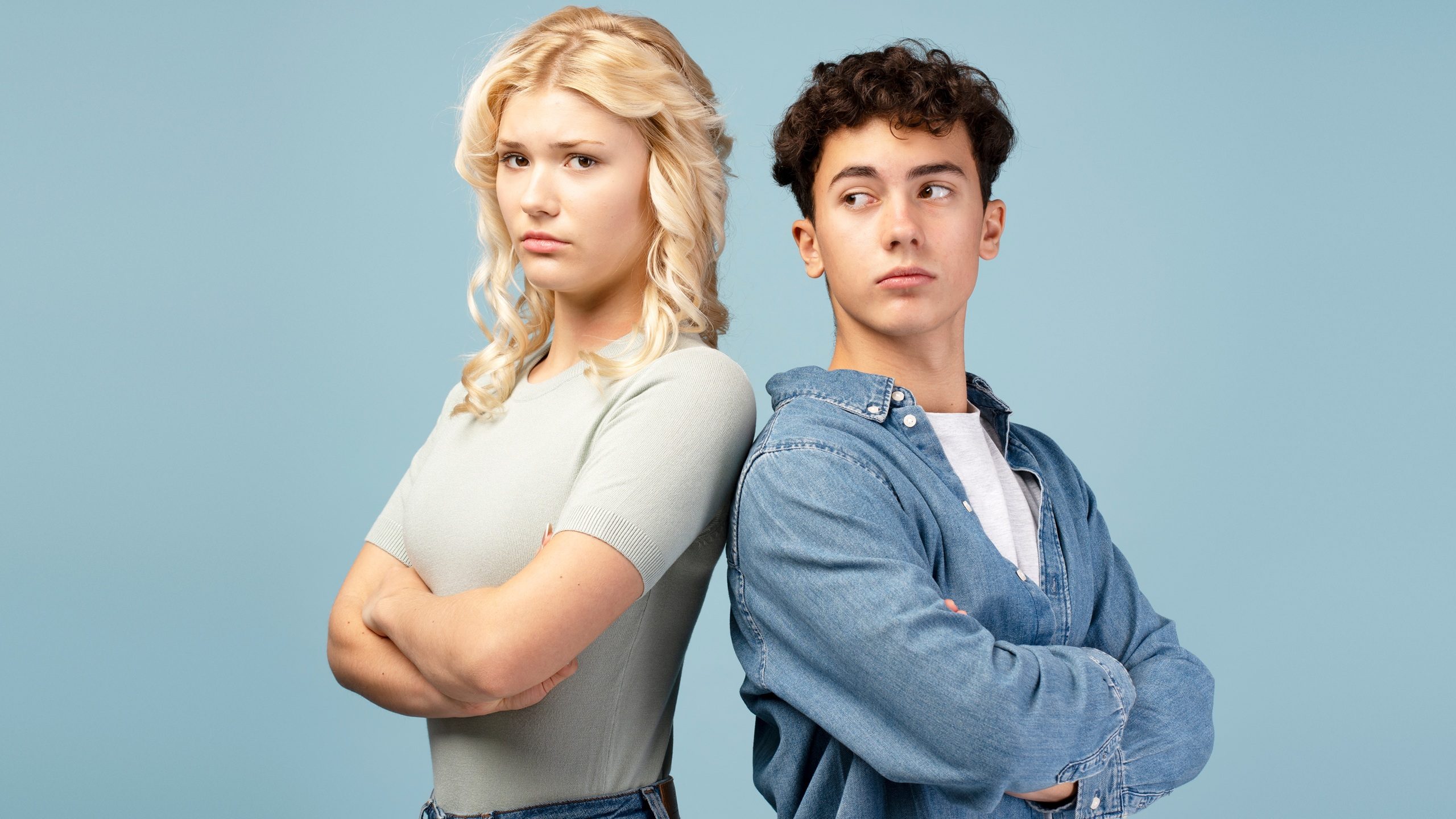 Upset quarreling couple, young guy and girl with crossed arms standing, back to back, looking away isolated on blue background. Conflict, relationship concept