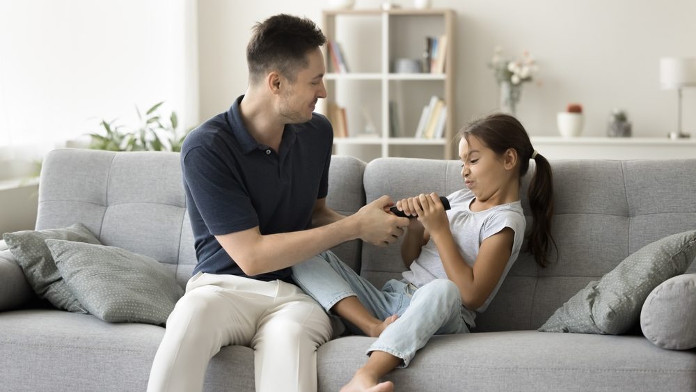 Serious father and little daughter sit on sofa grabbing, fighting for remote controller from tv, preschooler child want to see cartoons, while her dad desired for sport match. Addiction, disobedience