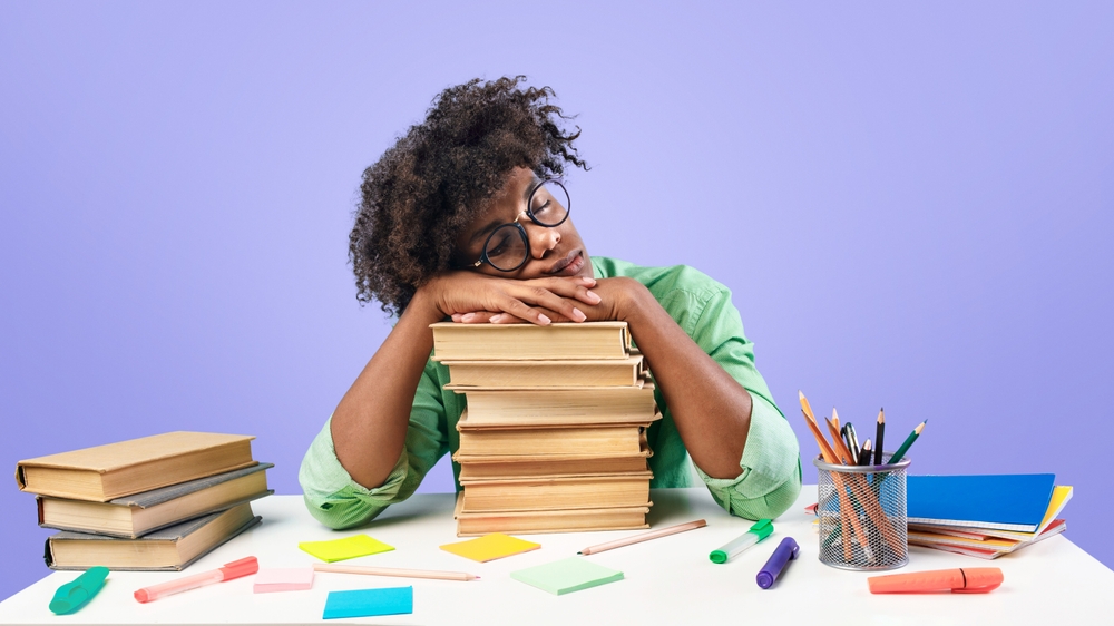 Exhausted black male student sleeping on desk on stacks of books, being tired while preparing for exam on blue studio background. African american teenager feeling overworked while studying