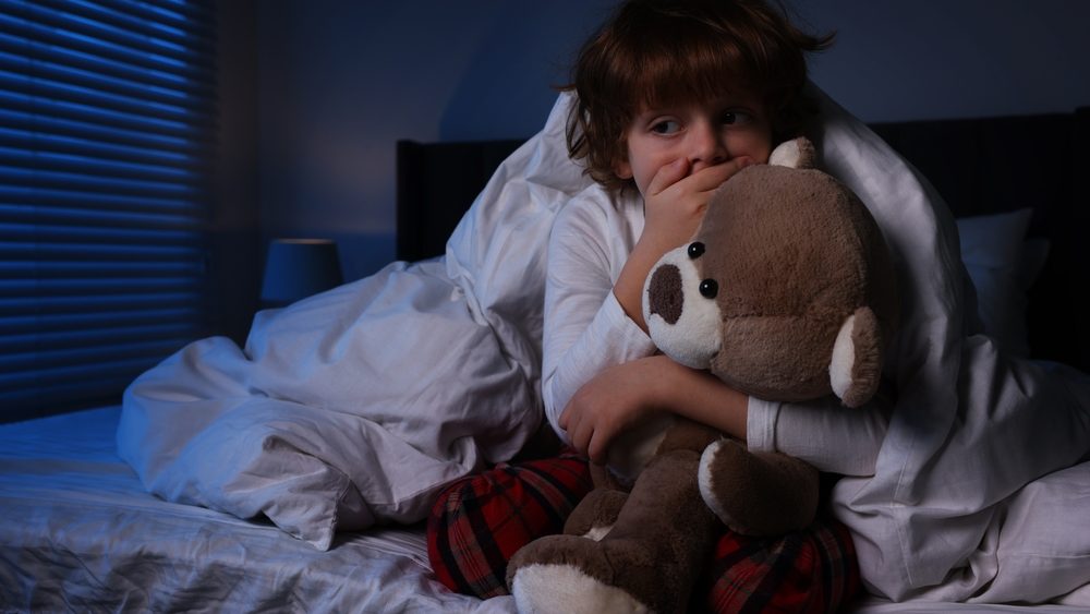 Scared boy with teddy bear in bed at night. Space for text