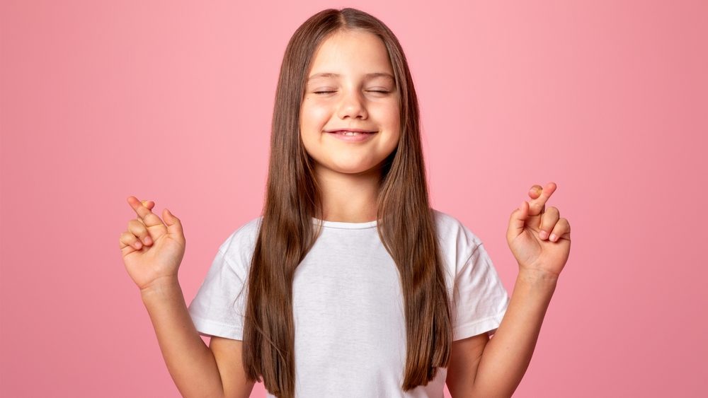 Waiting for surprise. Smiling child with closed eyes and crossed fingers makes wish, isolated on pink background, studio shoot