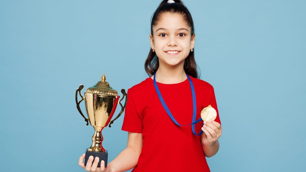 Little kid girl 8-9 years old she wears red t-shirt golden medal hold in hand champion cup have fun isolated on blue color background child studio portrait. Mother's Day love family lifestyle concept