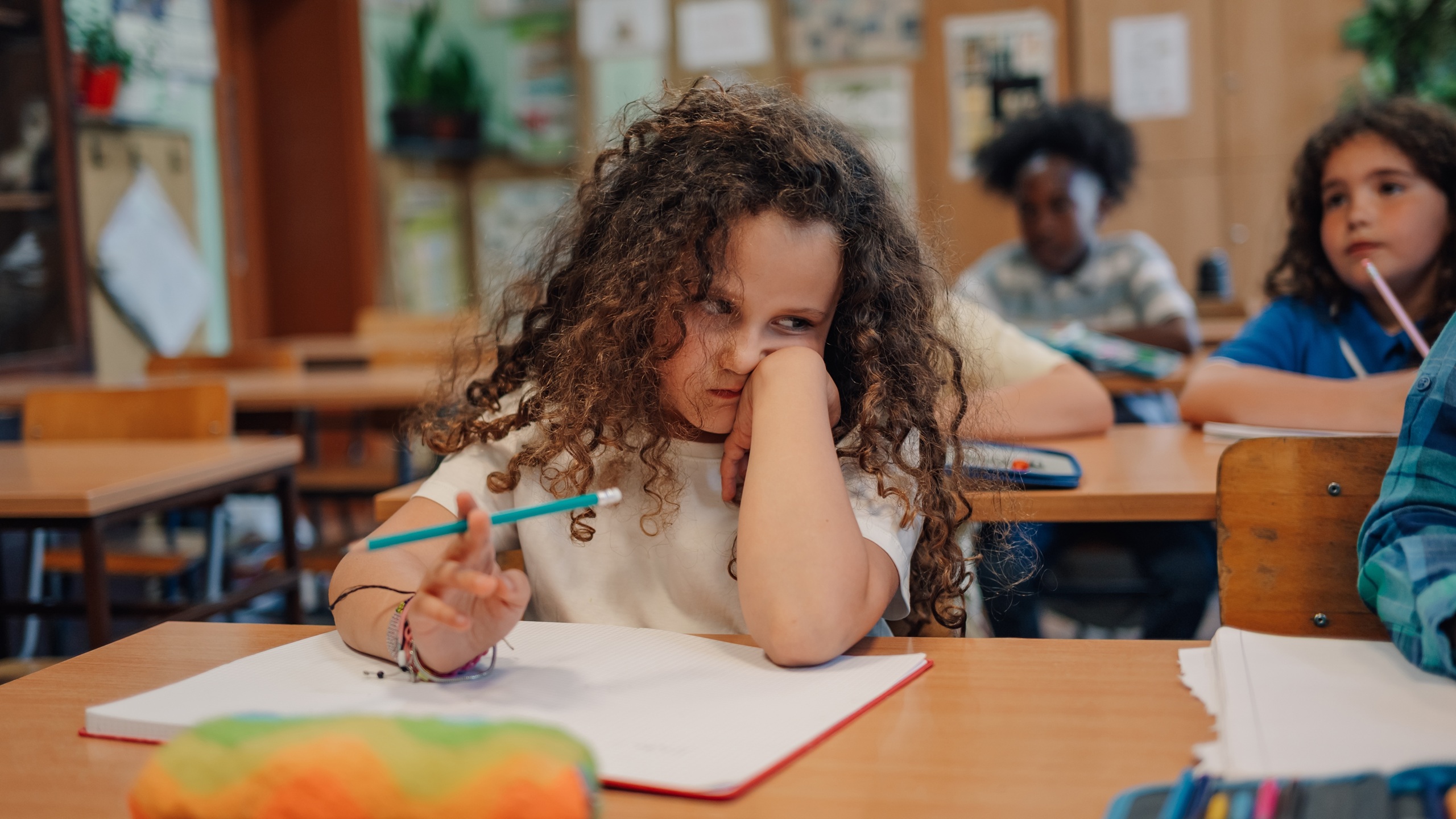 Bored elementary school student is sitting at desk and struggling with schoolwork while her classmates are working