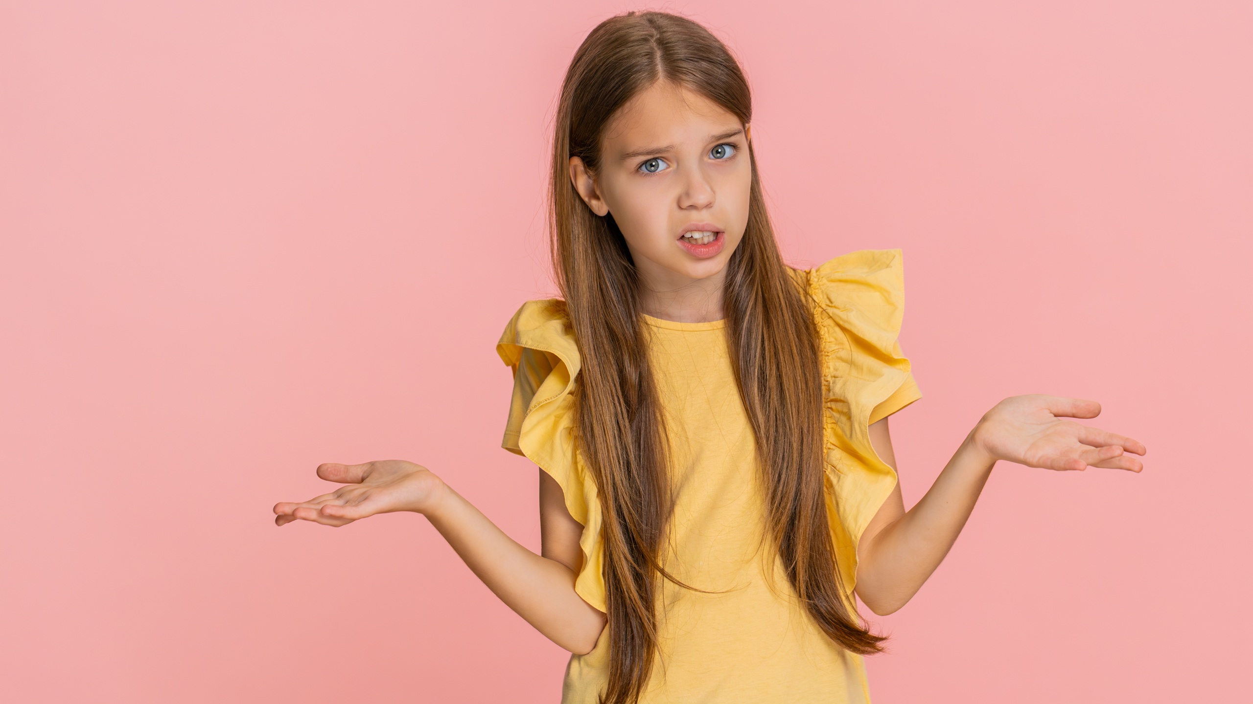 Young child girl raises eyebrows in confusion, showing hand gesture meaning what, why, expressing surprise and curiosity. School kid isolated on pink background with emotional and playful reaction.
