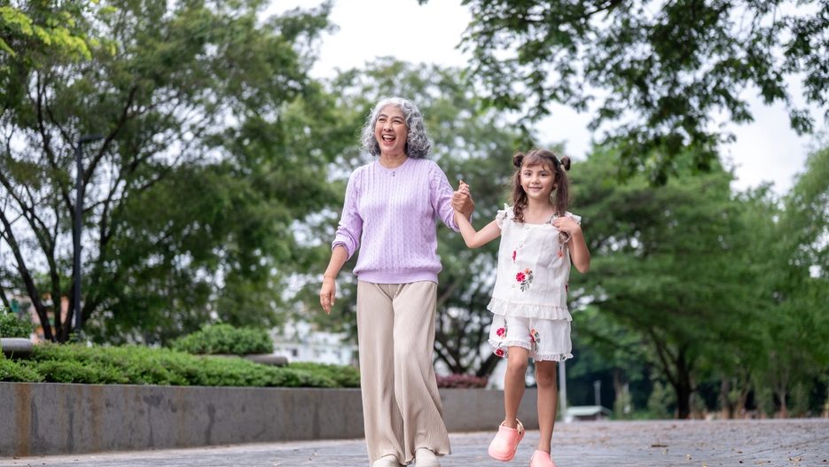 Smiling old woman grandma holding hand and walking with kid girl granddaughter on pavement in park. Child development, Public space, Outdoor Activity.