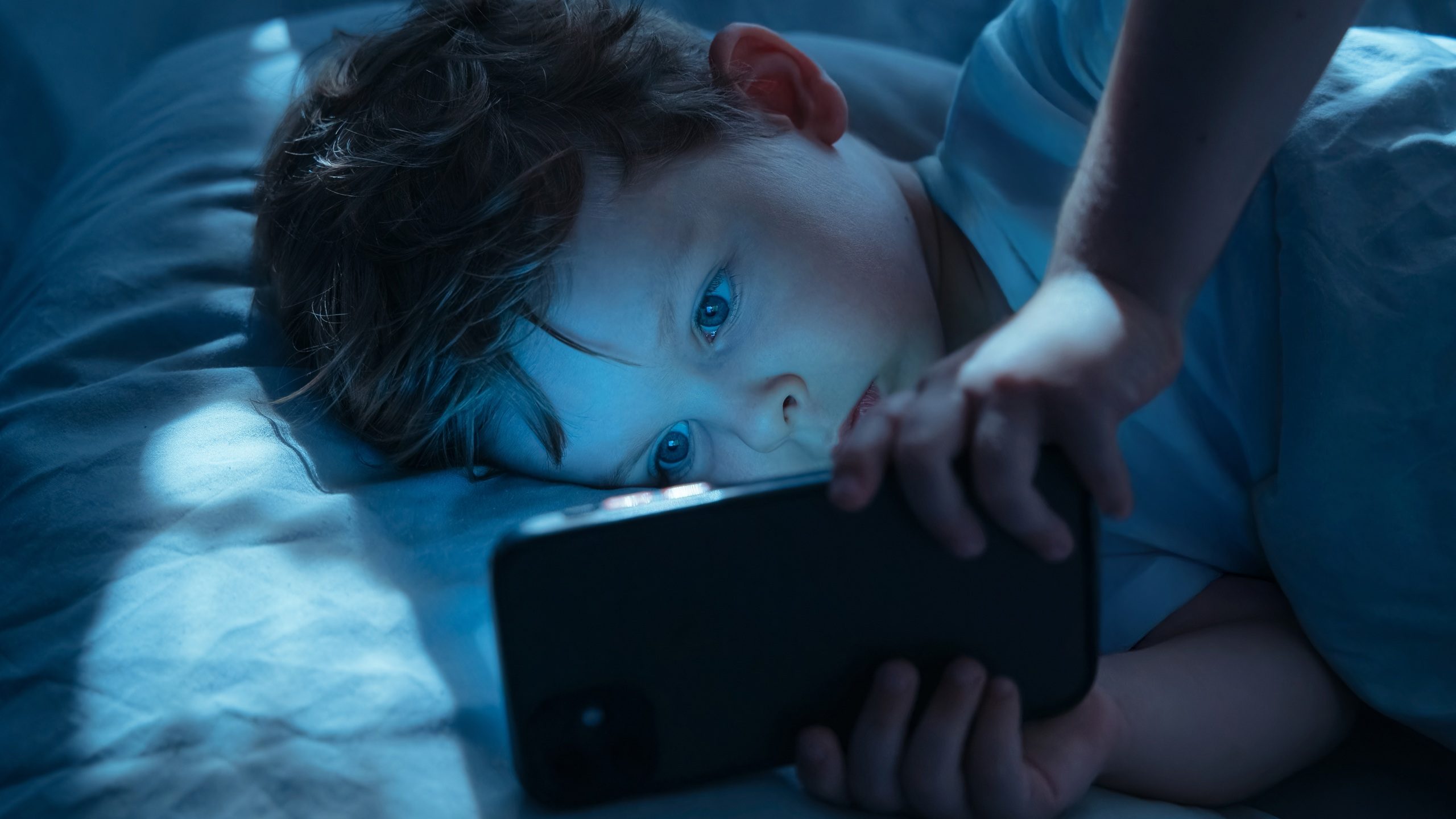 Little boy lies in bed at night, staring intently at his smartphone screen. Child watches cartoons on his gadget, illuminated by blue light of the screen. Childhood and technology, Digital addiction