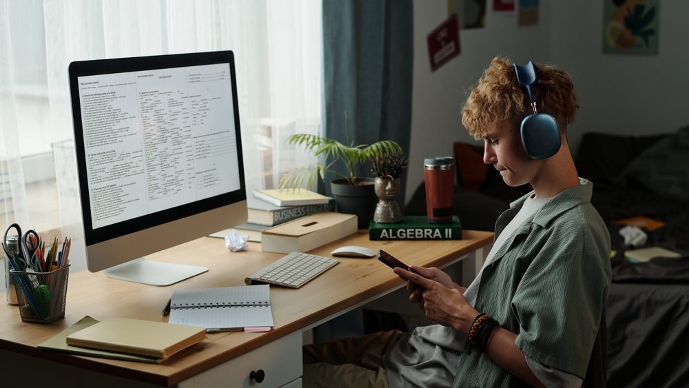 Caucasian teenage boy wearing headphones using smartphone while ignoring computer screen with study materials showing signs of gadget addiction in home bedroom setting