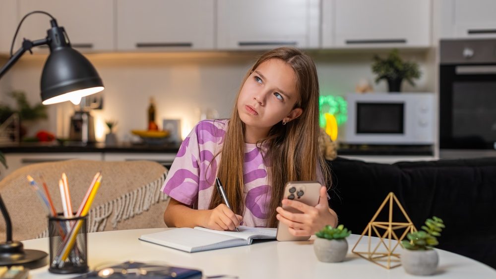 Teenager girl at night home watches online math lesson on smartphone makes notes in notebook. Caucasian child kid sitting at table at evening studies math writes formula solves tasks focus learning.